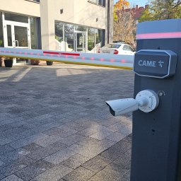 SPECJALISTA.NET SPÓŁKA Z OGRANICZONĄ ODPOWIEDZIALNOŚCIĄ - Szlaban automatyczny CAME z kamerą monitoringu na tle budynku. Widoczne czerwone światła LED na szlabanie i logo CAME na obudowie. Kostka brukowa na podłożu.