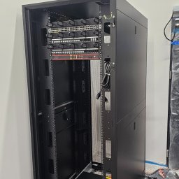 SPECJALISTA.NET SPÓŁKA Z OGRANICZONĄ ODPOWIEDZIALNOŚCIĄ - Częściowo wypełniona szafa rack z zamontowanymi przełącznikami sieciowymi, okablowaniem i zasilaczem UPS, widoczne kable w korytku.