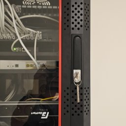 SPECJALISTA.NET SPÓŁKA Z OGRANICZONĄ ODPOWIEDZIALNOŚCIĄ - Zamknięta szafa rack z kluczykiem w zamku, widoczne kable i urządzenia sieciowe wewnątrz. Czerwona listwa ozdobna. Widok z boku, oświetlenie dzienne.