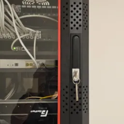 Zamknięta szafa rack z kluczykiem w zamku, widoczne kable i urządzenia sieciowe wewnątrz. Czerwona listwa ozdobna. Widok z boku, oświetlenie dzienne.