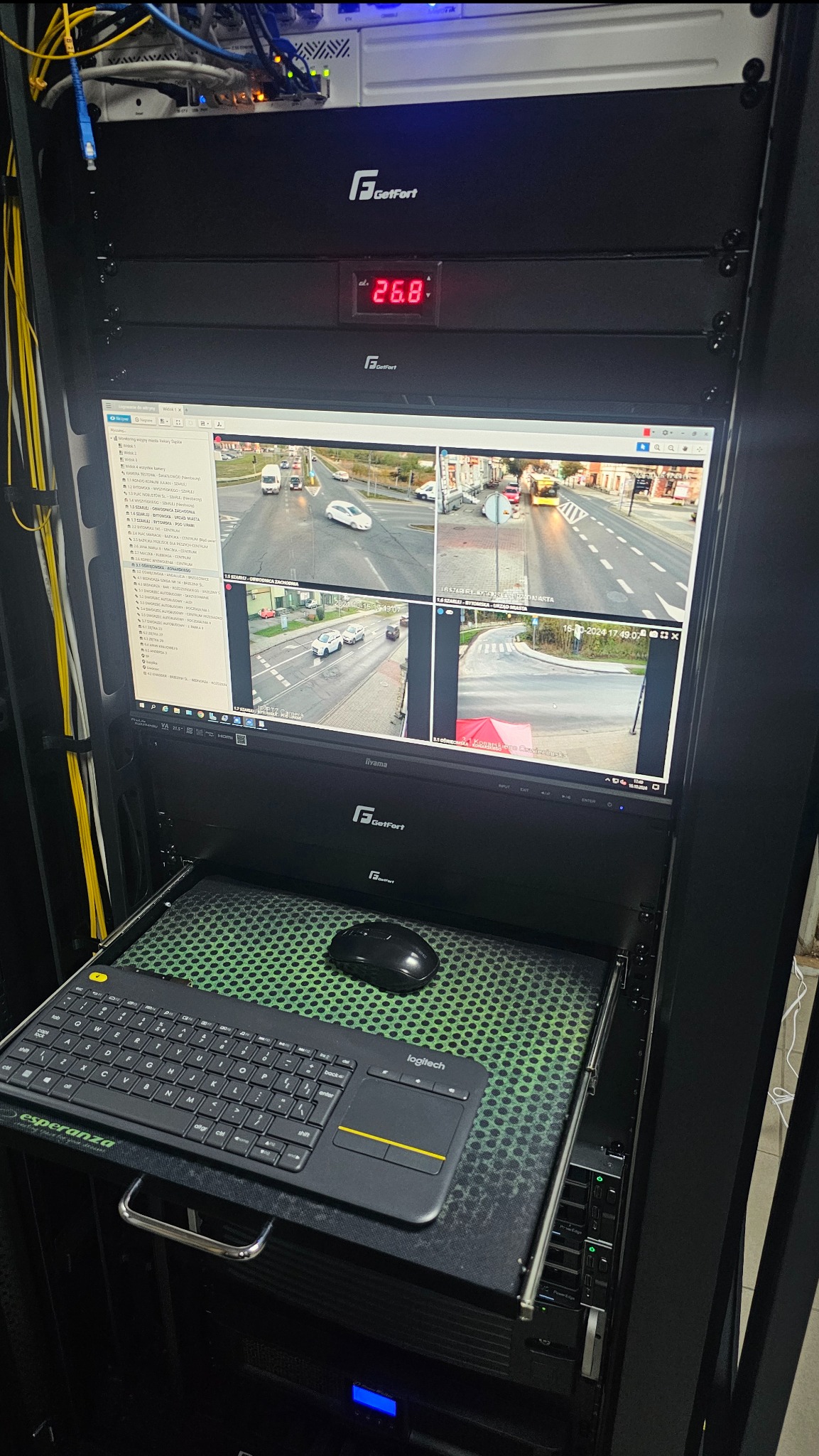Serwerownia z monitoringiem wizyjnym GetFort: ekran z podglądem z kamer na skrzyżowaniu, klawiatura Logitech, myszka. Widoczne kable i urządzenia sieciowe.