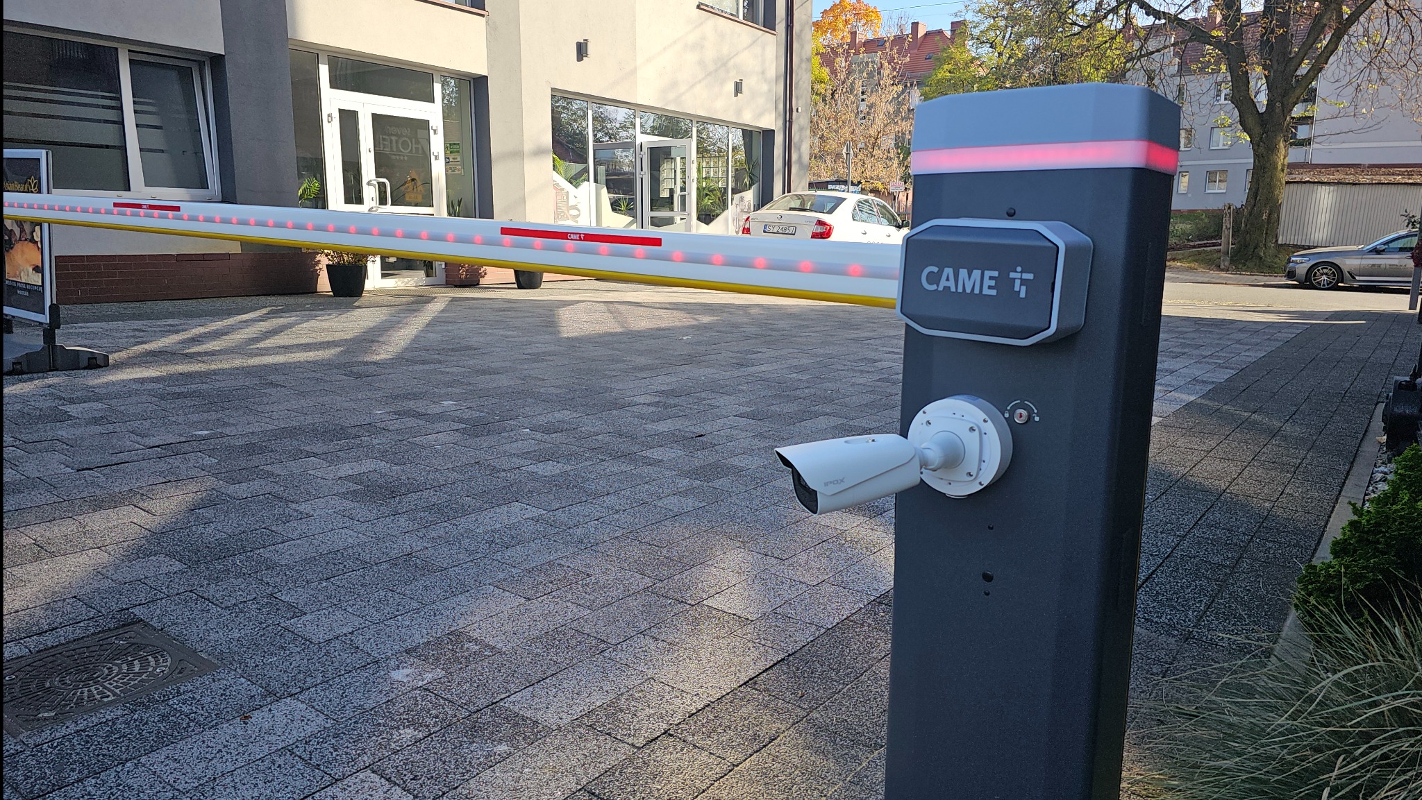 Szlaban automatyczny CAME z kamerą monitoringu na tle budynku. Widoczne czerwone światła LED na szlabanie i logo CAME na obudowie. Kostka brukowa na podłożu.