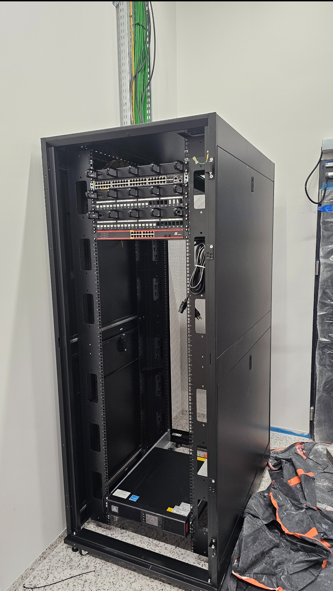 Częściowo wypełniona szafa rack z zamontowanymi przełącznikami sieciowymi, okablowaniem i zasilaczem UPS, widoczne kable w korytku.