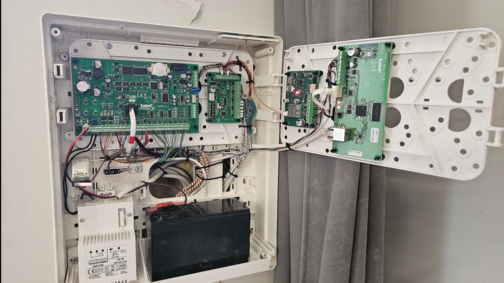 Otwarta obudowa centrali alarmowej Satel Integra 32 z widocznymi płytkami drukowanymi, przewodami i akumulatorem. Widoczne transformator AWT150. Szara ściana w tle.