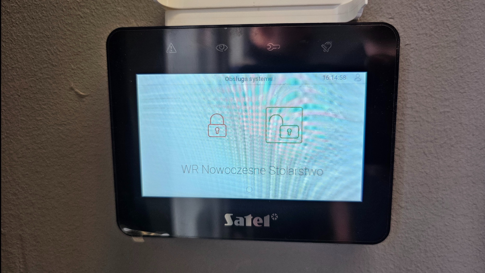 Panel dotykowy centrali alarmowej Satel na ścianie, prezentujący stan systemu z ikonami kłódek, alarmu, oka i dzwonka. Widoczny napis 'WR Nowoczesne Stolarstwo'.