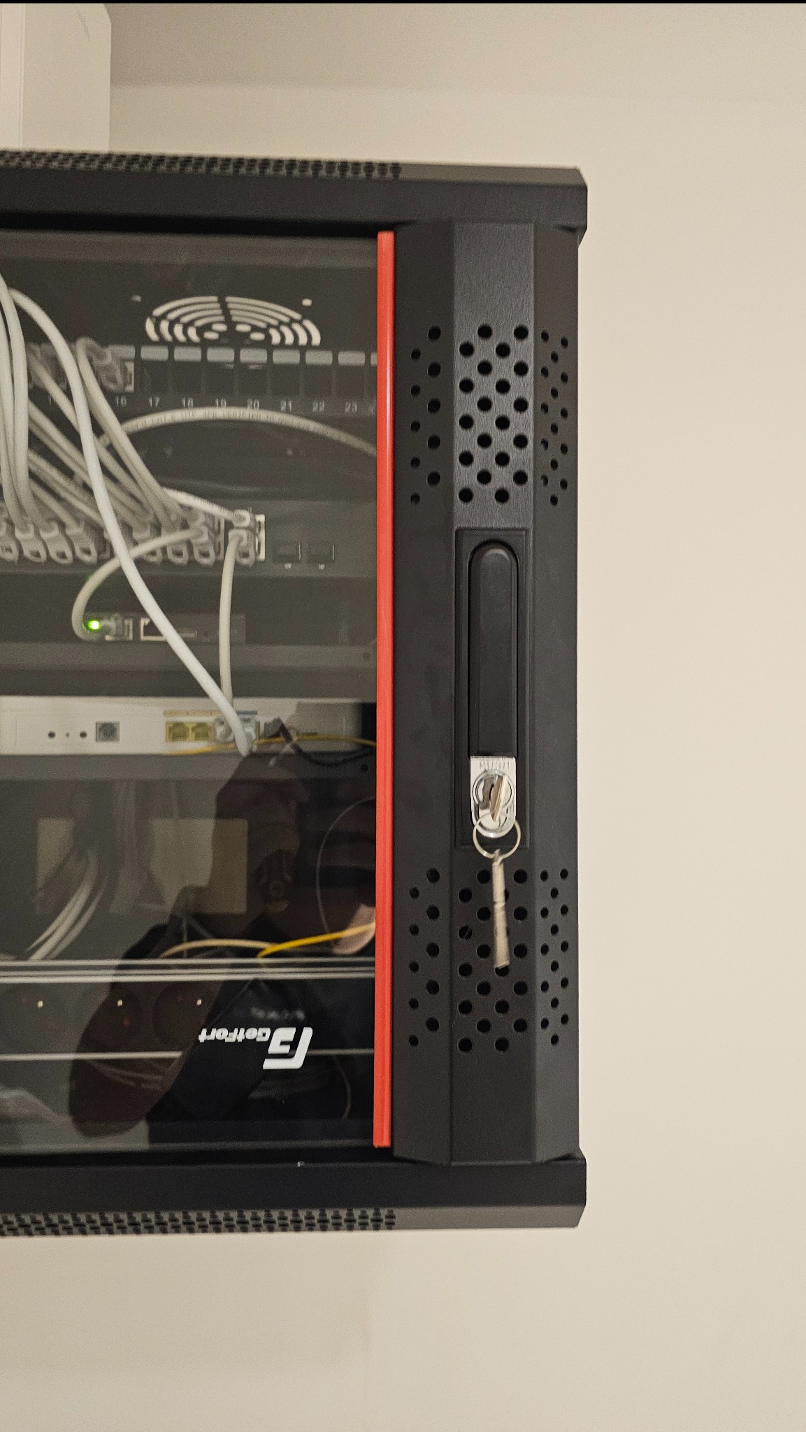 Zamknięta szafa rack z kluczykiem w zamku, widoczne kable i urządzenia sieciowe wewnątrz. Czerwona listwa ozdobna. Widok z boku, oświetlenie dzienne.