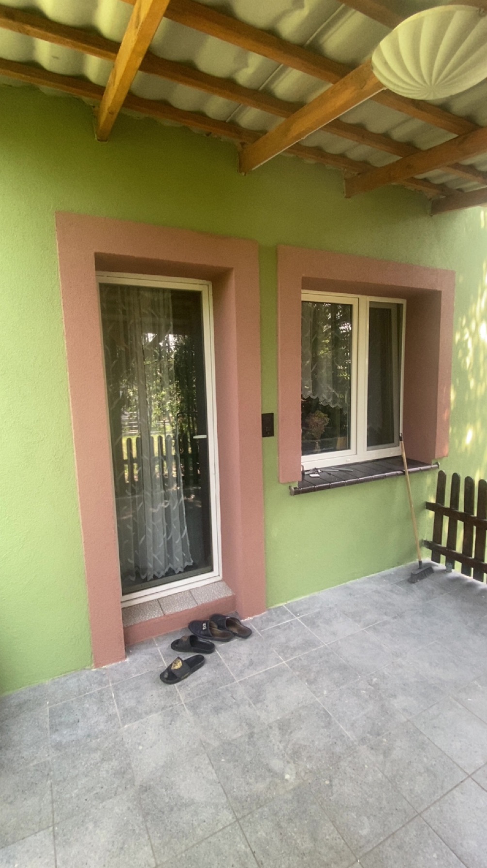 Ściana domu pomalowana na zielono z oknem i drzwiami w różowych obramowaniach. Na podłodze leżą klapki. Widoczny fragment dachu i lampa.