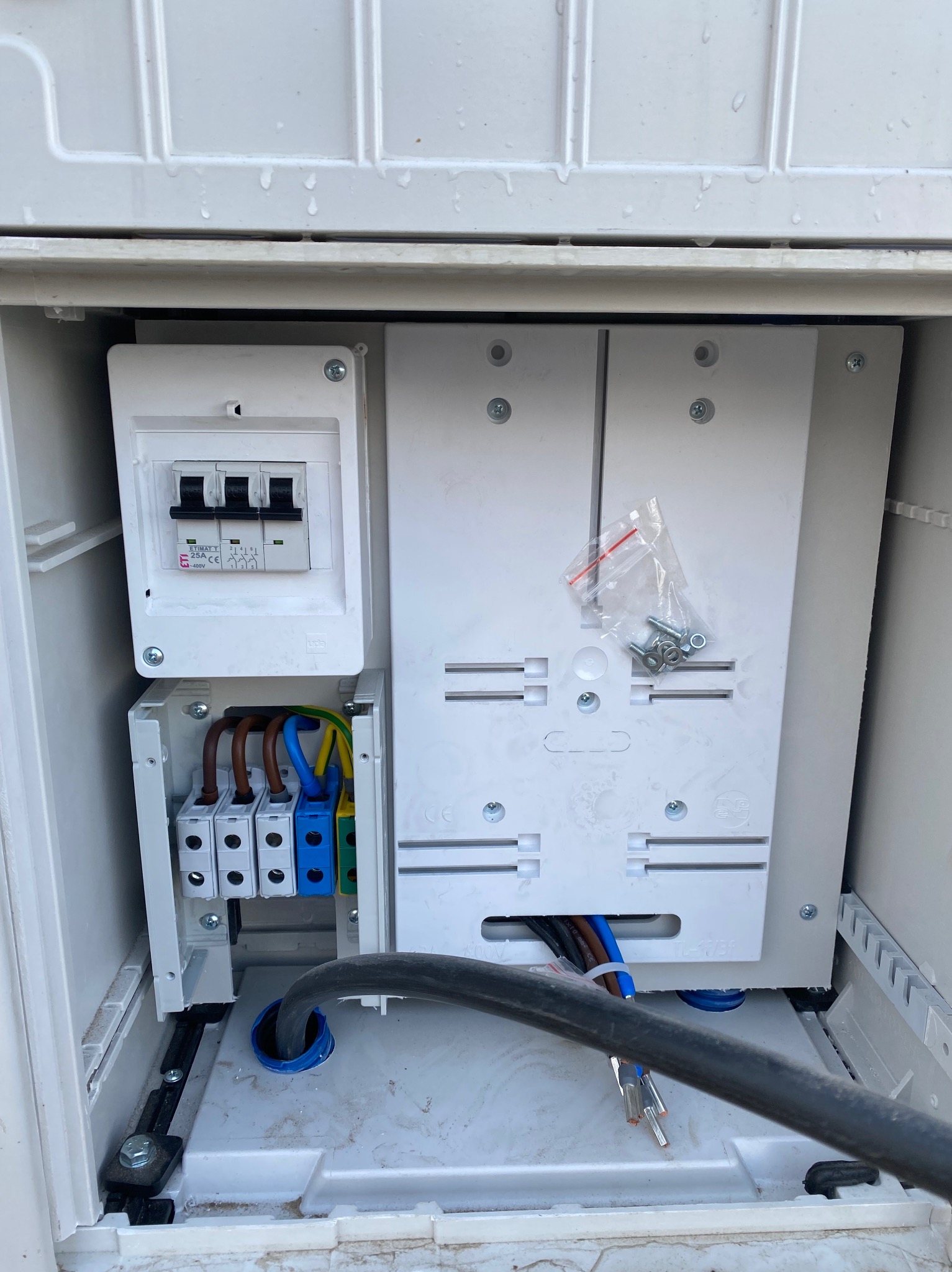 Otwarta, biała skrzynka elektryczna z zabezpieczeniami, przewodami i elementami montażowymi. Widoczne bezpieczniki, złączki, kable w kolorach: brązowy, niebieski, żółto-zielony.