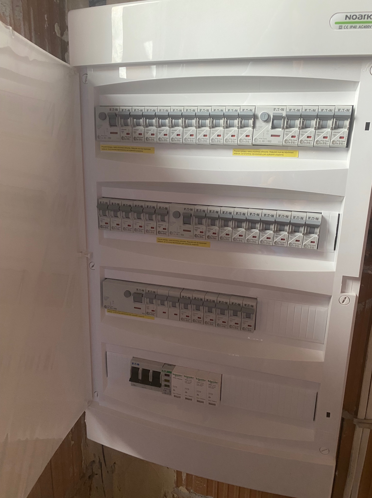 Otwarta, biała skrzynka rozdzielcza z zainstalowanymi bezpiecznikami EATON i Schneider, widoczna na tle surowej ściany. Instalacja elektryczna w trakcie prac.