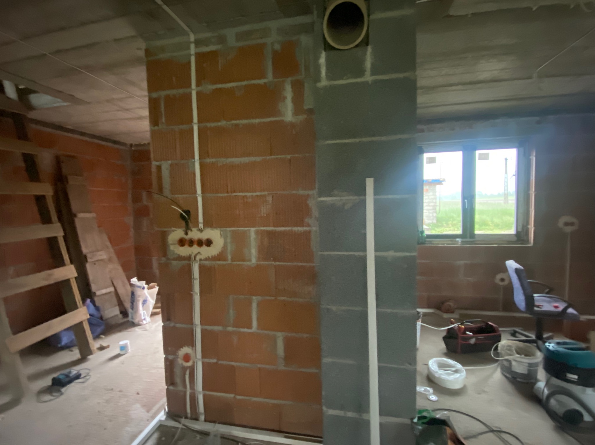 Wnętrze w trakcie budowy: widoczne ceglane ściany z instalacją elektryczną, fragment okna i narzędzia, ukazujące etap prac wykończeniowych.