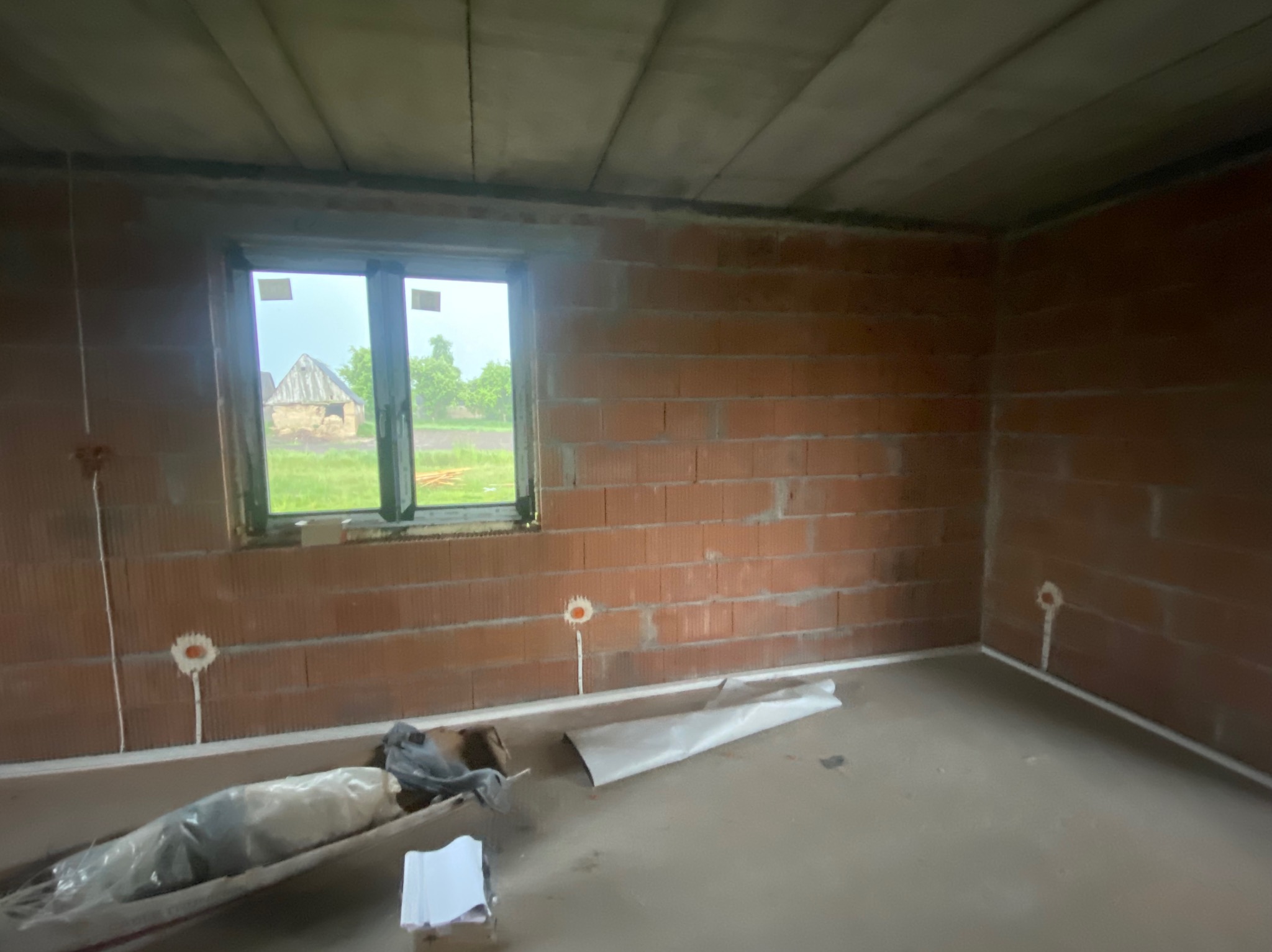 Surowy stan wnętrza domu w budowie: ściany z cegły, okno z widokiem na krajobraz, widoczne instalacje elektryczne, betonowa podłoga i sufit.