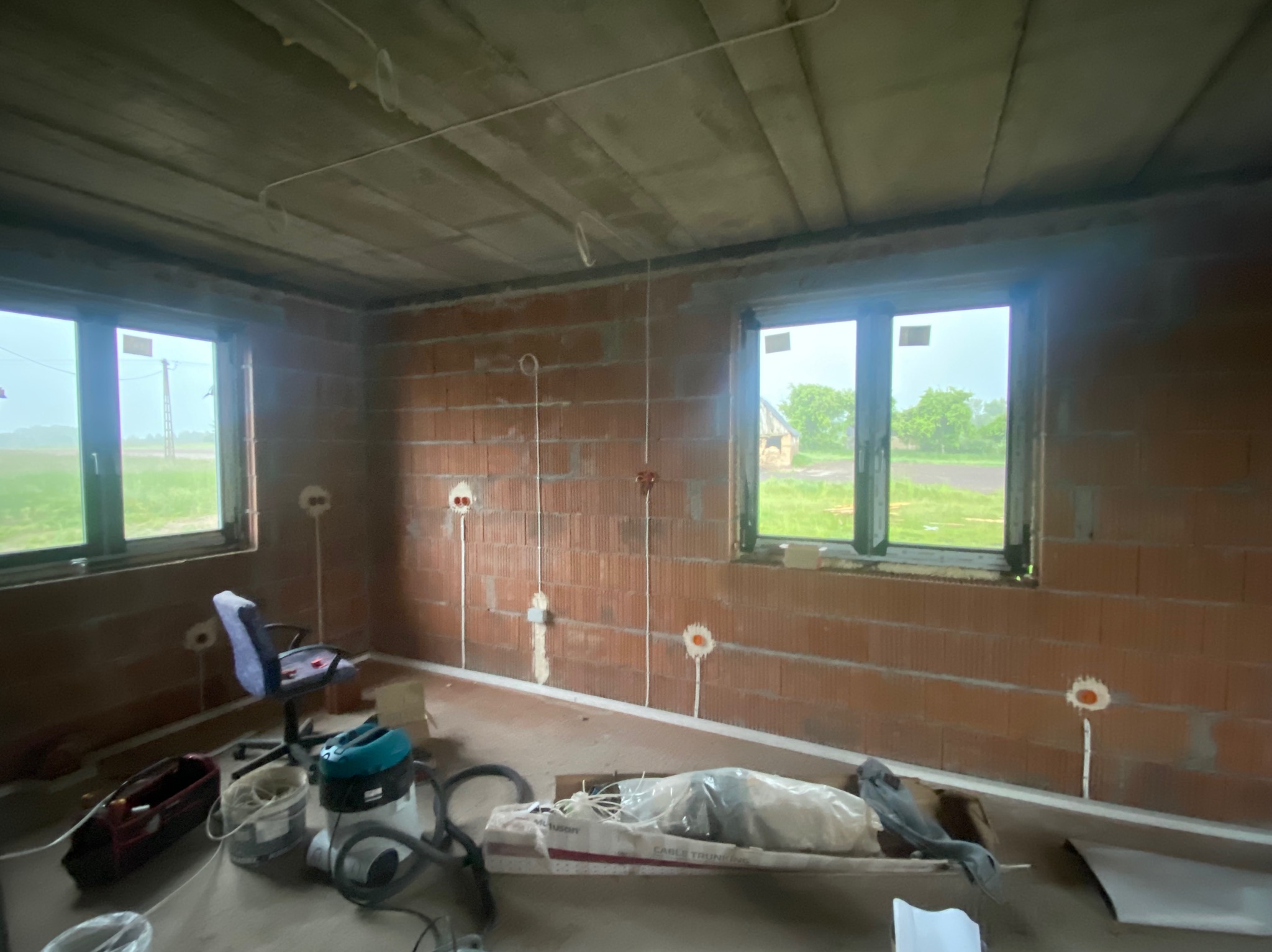 Wnętrze surowego domu z cegły z widocznymi puszkami elektrycznymi i przewodami. Dwa okna z widokiem na zewnątrz. Na podłodze narzędzia i materiały budowlane.