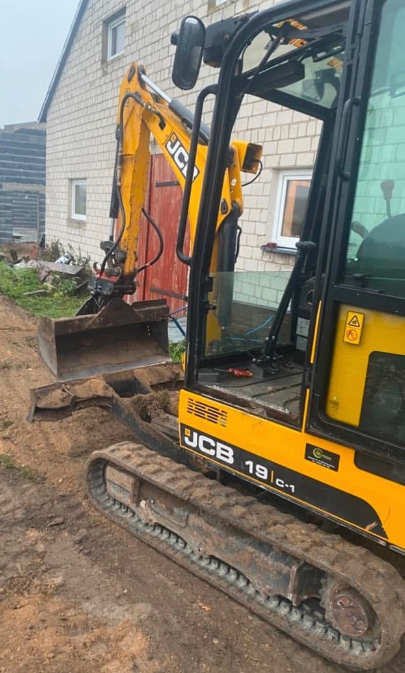 Żółta mini koparka JCB 19C-1 na placu budowy, z uniesioną łyżką. W tle widoczny fragment budynku z jasnej cegły i częściowo wykopana ziemia.