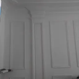 Biała ściana z dekoracyjnymi listwami i sztukaterią, widoczne elementy urządzenia na półce i fragment krzesła w tle.