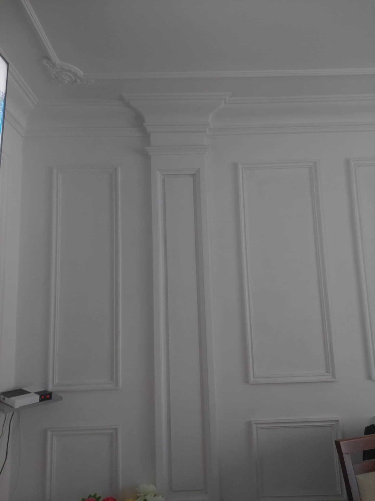 Biała ściana z dekoracyjnymi listwami i sztukaterią, widoczne elementy urządzenia na półce i fragment krzesła w tle.