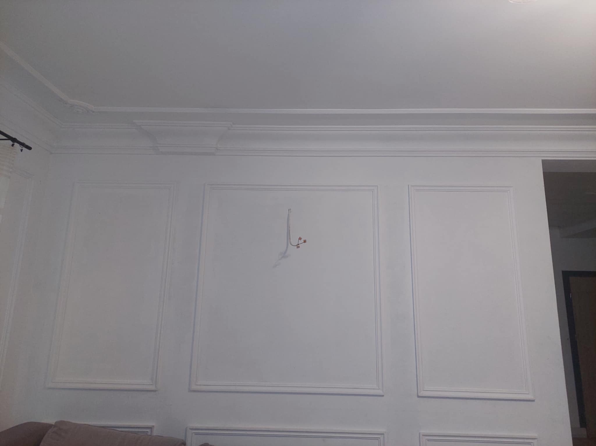 Biała ściana z ozdobnymi panelami i listwami przypodłogowymi, widoczne przewody elektryczne wystające ze ściany, fragment sofy w dolnej części kadru.