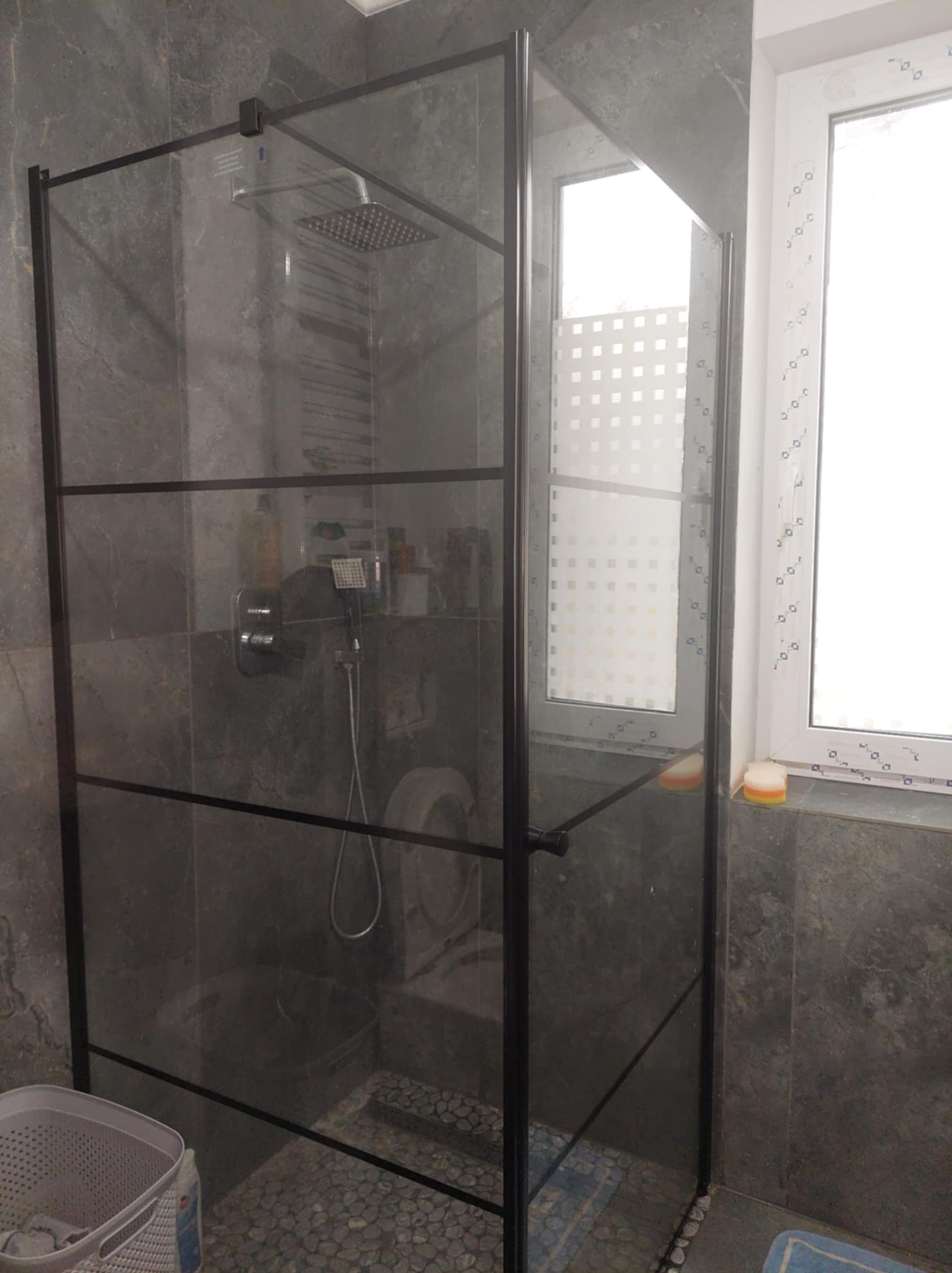 Kabina prysznicowa z czarnymi profilami na tle szarych płytek imitujących kamień, widoczne okno z ozdobną folią, odpływ liniowy i kosz na pranie.