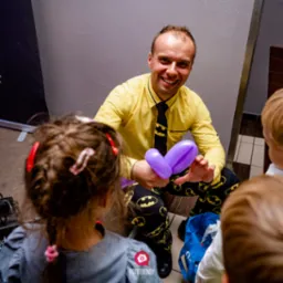 Animator w żółtej koszuli i spodniach z motywem Batmana, uśmiechnięty, modeluje baloniki dla dzieci na imprezie.