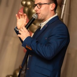 Dj Blażejos - Dj na wesele Imprezy okolicznościowe i firmowe - Mężczyzna w niebieskim garniturze, okularach i z mikrofonem w dłoni, gestykuluje podczas występu na tle dekoracji z balonów.