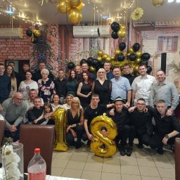 Dj Blażejos - Dj na wesele Imprezy okolicznościowe i firmowe - Duża grupa osób pozuje do zdjęcia w udekorowanym lokalu podczas osiemnastych urodzin, widoczne złote i czarne balony, dekoracje świetlne i fototapeta z włoskim miasteczkiem.