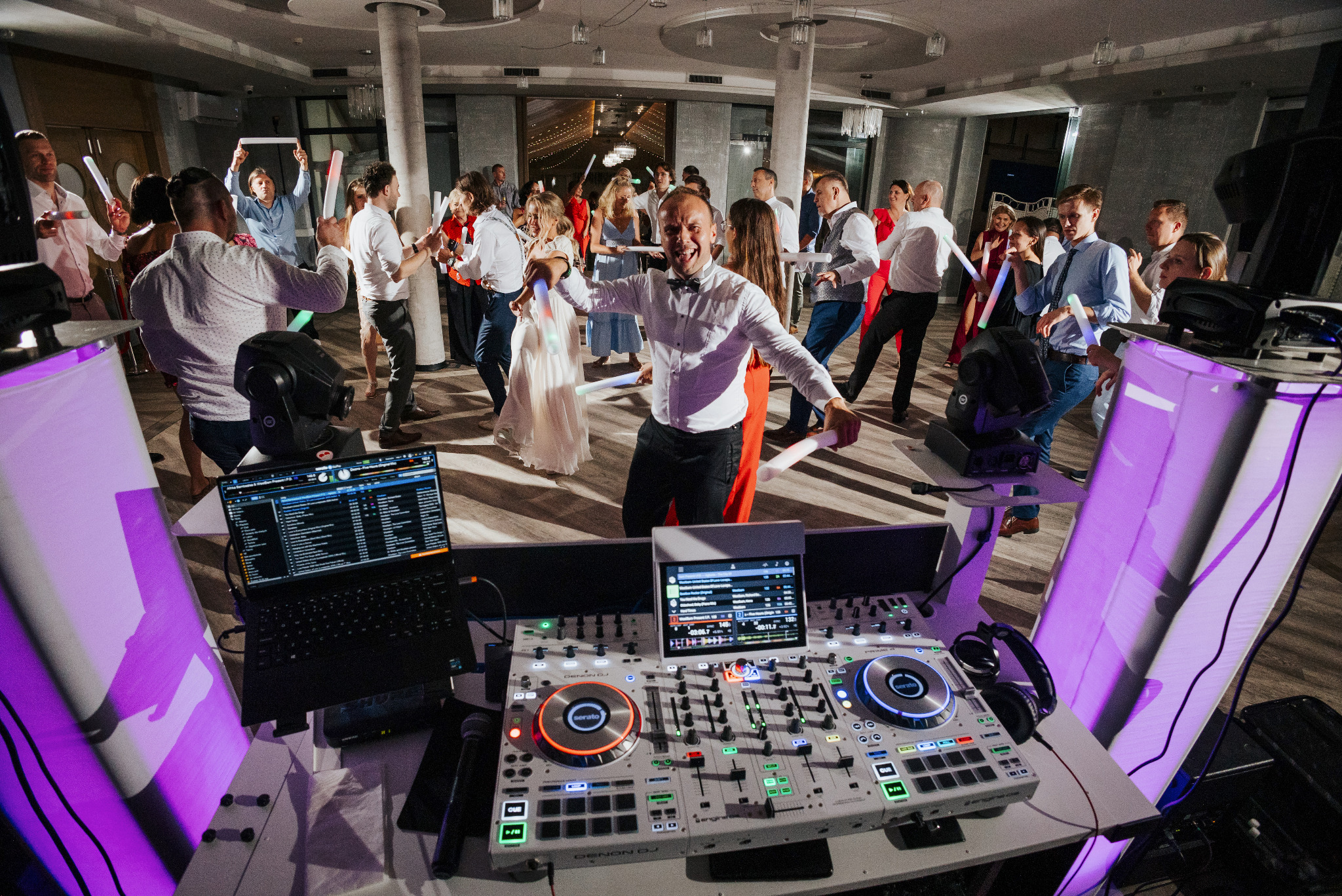 Zespół muzyczny na weselu, DJ tańczy z gośćmi trzymając świetlne pałeczki, widoczny sprzęt DJ-ski z podświetleniem.