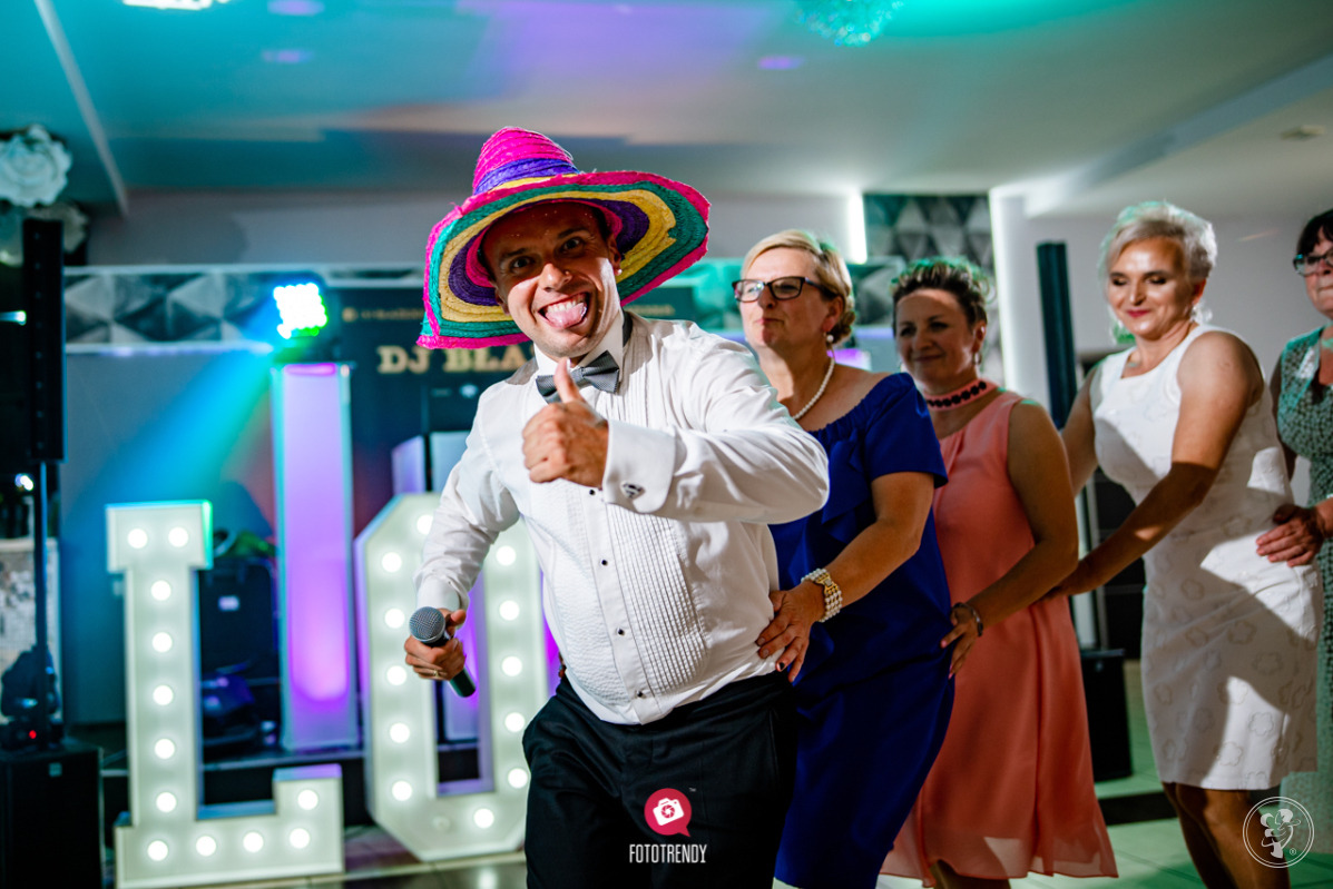 Mężczyzna w kolorowym sombrero i muszce, z mikrofonem w dłoni, pokazuje kciuk w górę, otoczony przez kobiety podczas zabawy weselnej, w tle podświetlane litery tworzące napis.