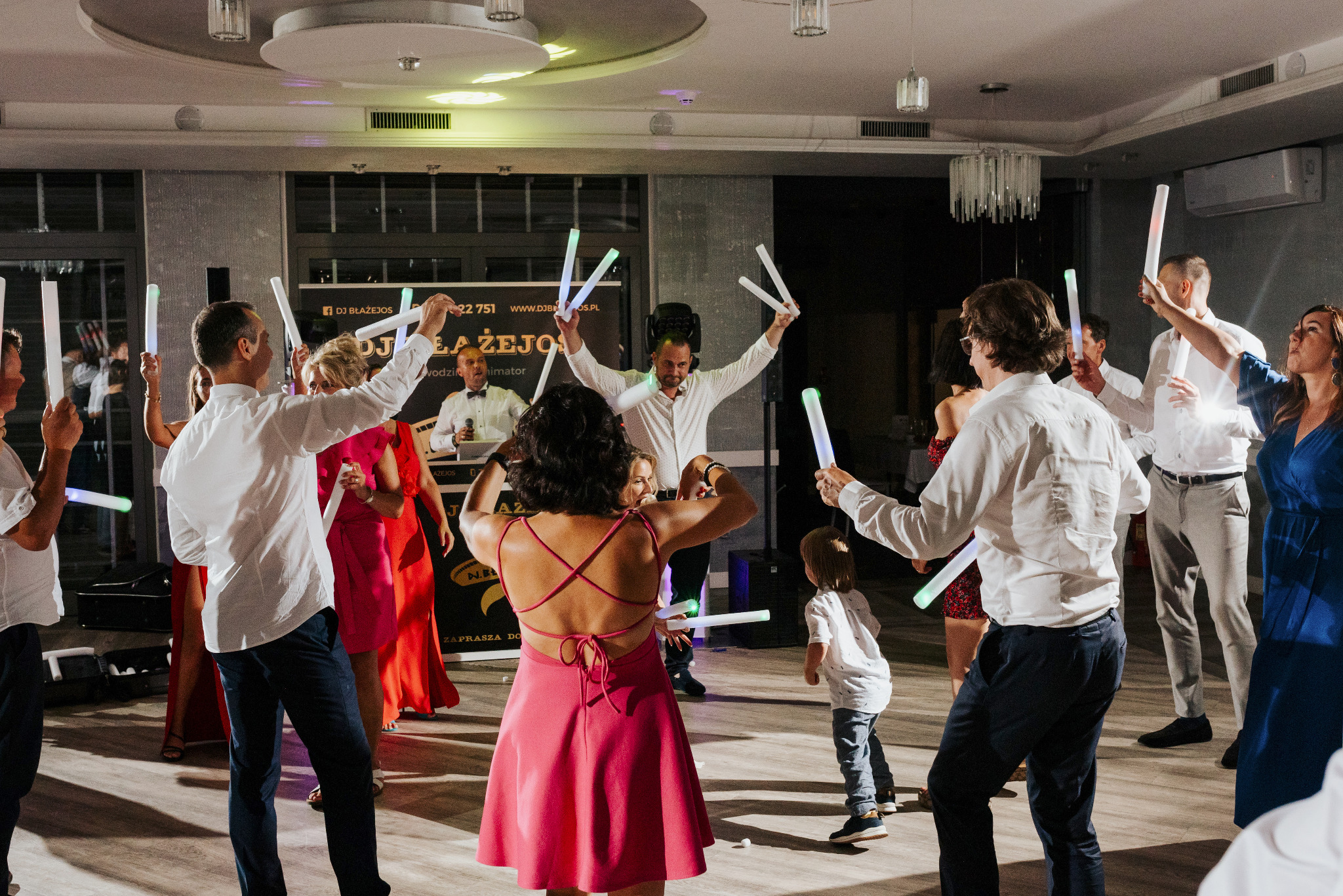 Rozbawieni goście weselni tańczą z ledowymi pałeczkami, wodzirej w tle za konsoletą DJ-ską.