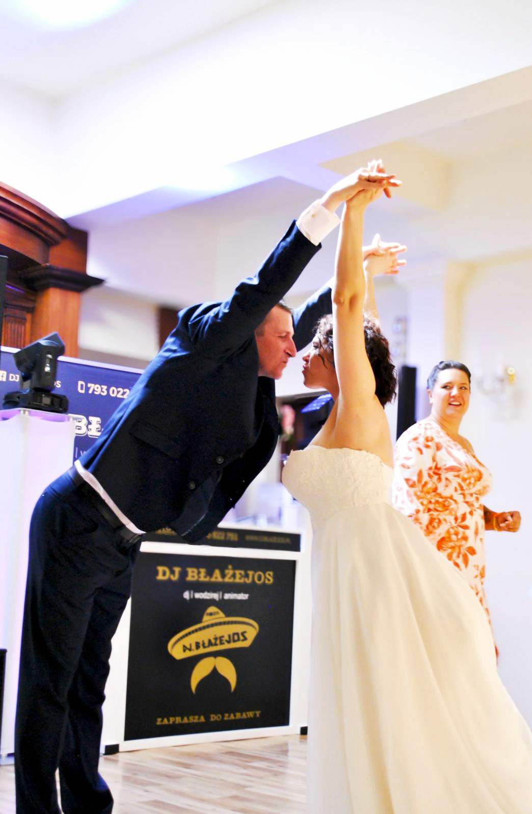 Panna młoda w białej sukni tańczy pod ramieniem mężczyzny w garniturze na tle stanowiska DJ z logo DJ Błażejos podczas wesela.