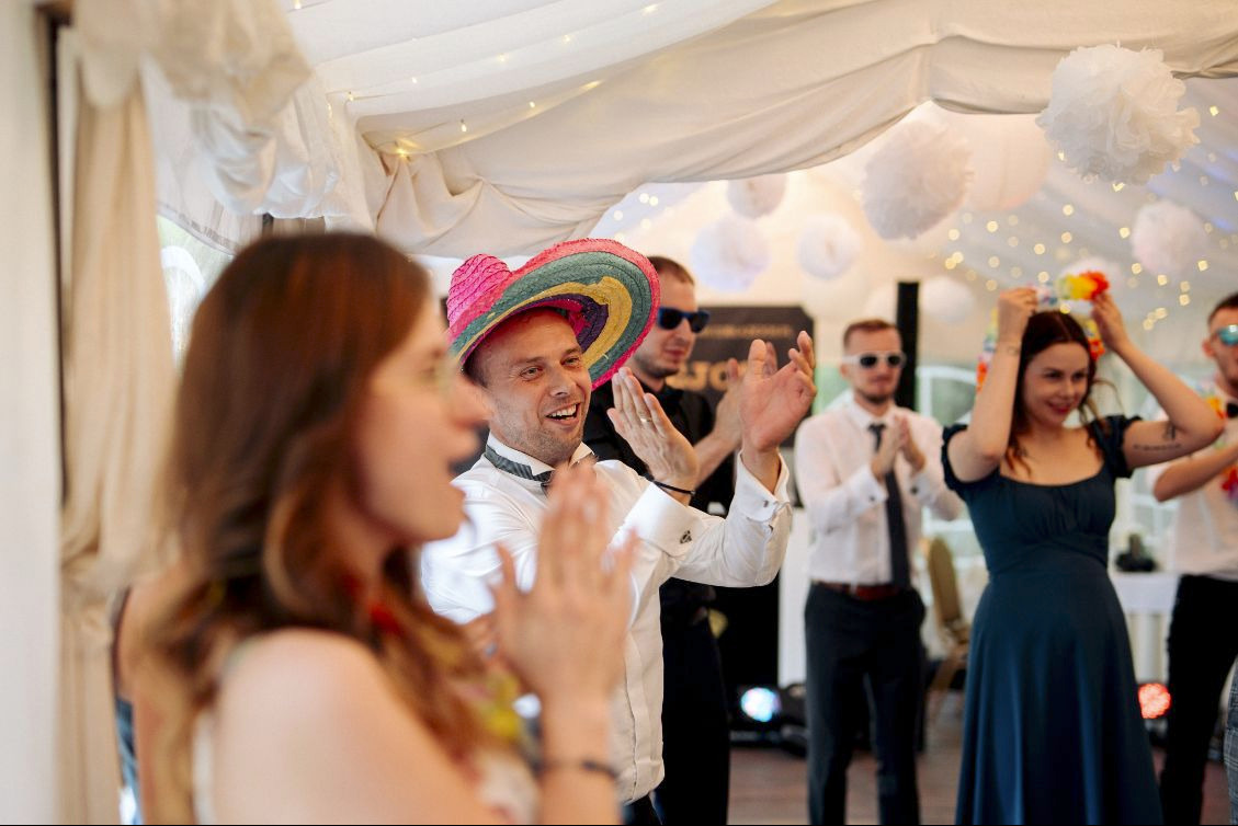 Uczestnicy imprezy w radosnych nastrojach, mężczyzna w barwnym sombrero w centrum uwagi, pod sufitem dekoracje z białych kul.