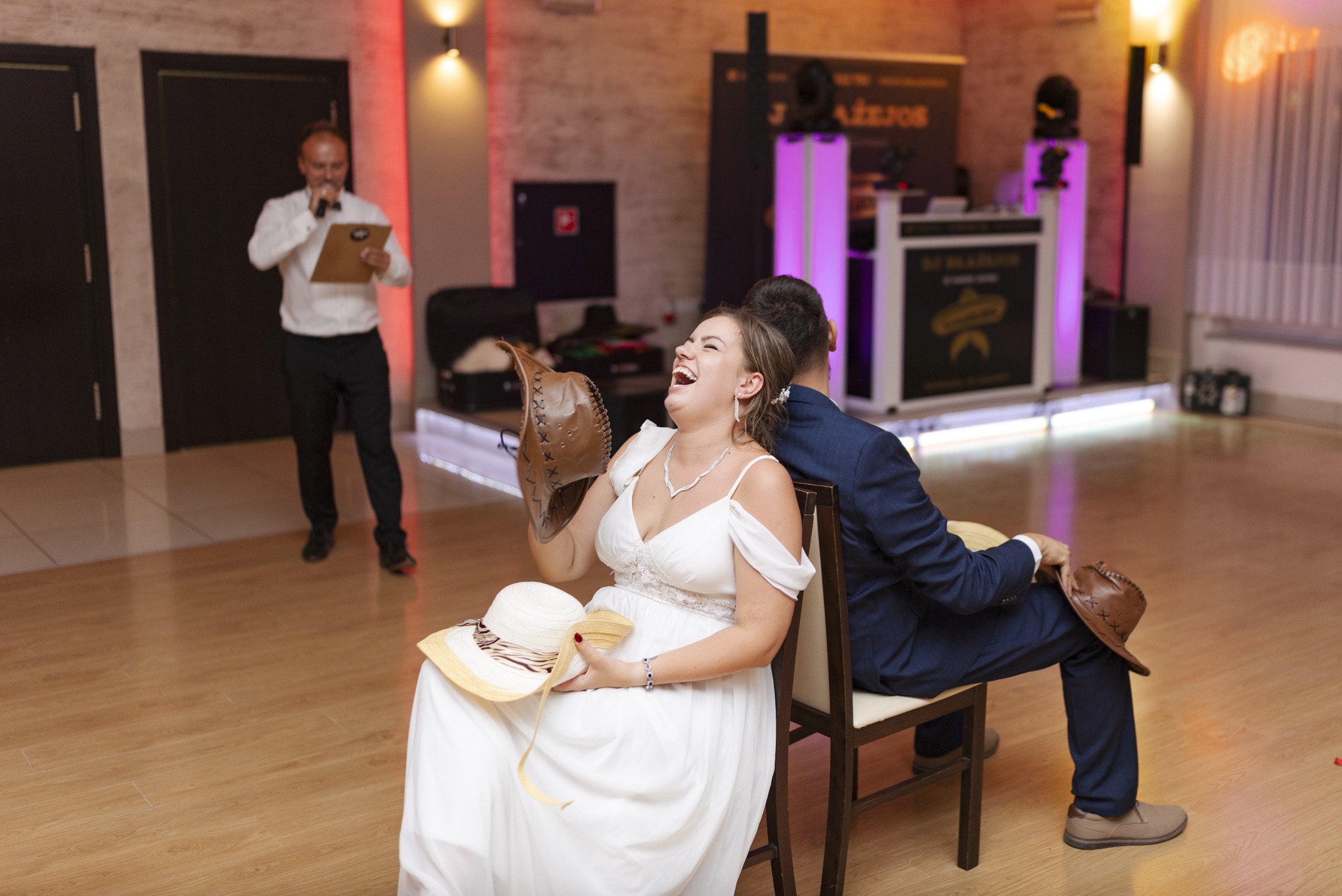 Panna młoda w białej sukni śmieje się, trzymając kapelusz kowbojski, podczas weselnej zabawy z quizem, gdzie para siedzi tyłem do siebie na krzesłach, a konferansjer w tle odczytuje pytania.