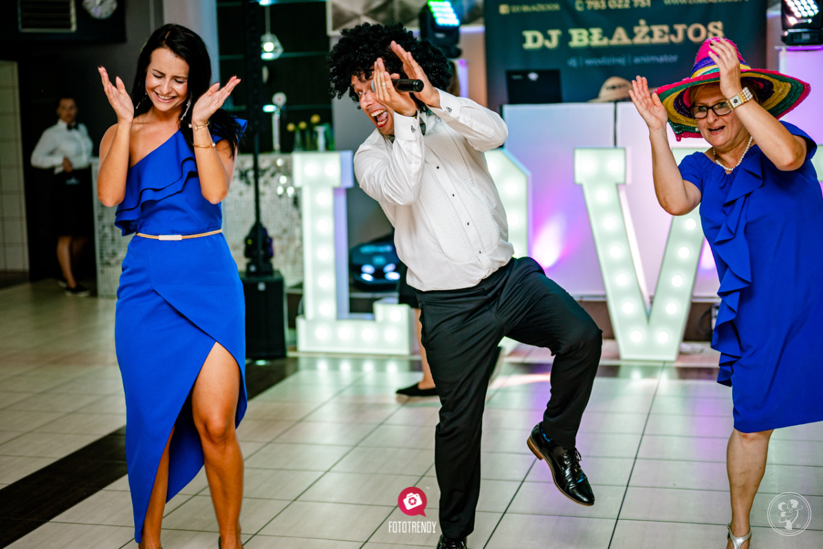 Rozbawiony konferansjer z afro peruką trzyma mikrofon, obok dwie kobiety w niebieskich sukienkach i z podniesionymi rękami, w tle podświetlony napis LOVE oraz DJ na podwyższeniu.