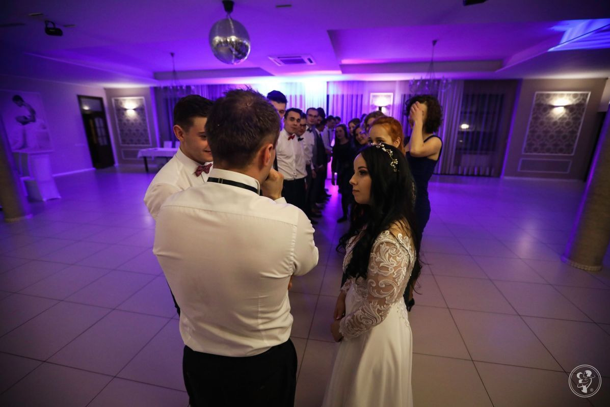 Para młoda w otoczeniu gości weselnych, oświetlenie sali w odcieniach fioletu, dekoracyjna kula dyskotekowa pod sufitem.