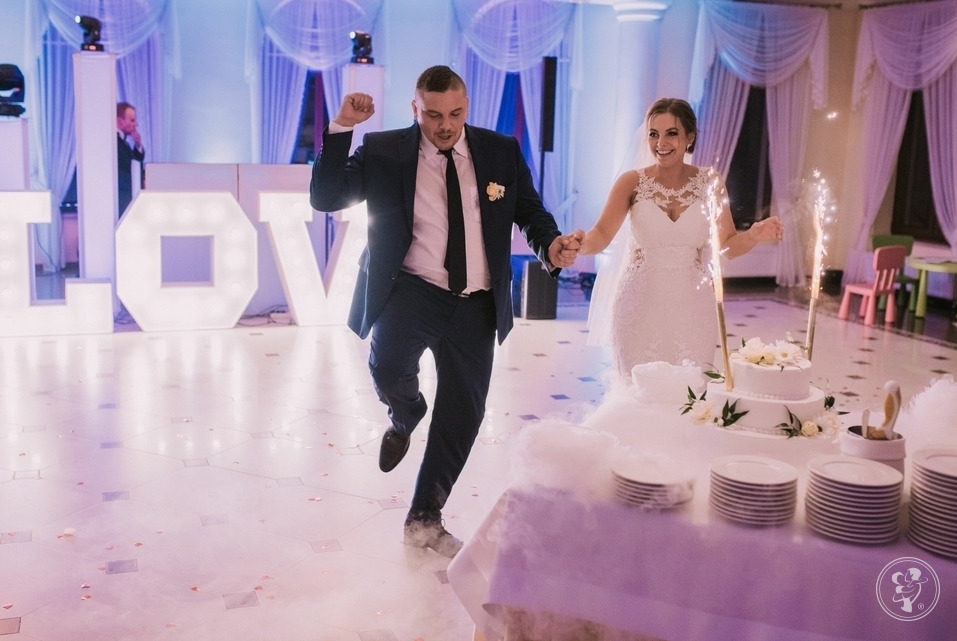 Uśmiechnięta para młoda, trzymająca się za ręce, w dynamicznym ruchu na sali weselnej z dekoracjami świetlnymi 'LOVE' i tortem z fontannami iskier.