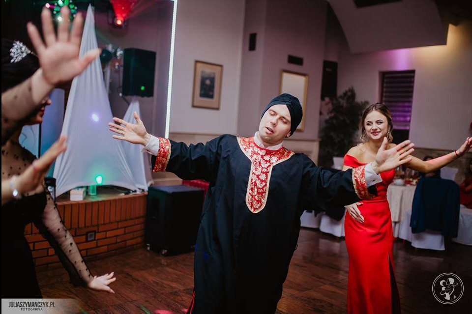 Występujący mężczyzna w czarnym stroju z czerwono-złotymi zdobieniami i turbanie podczas imprezy, w tle tańczące kobiety w sukniach wieczorowych.