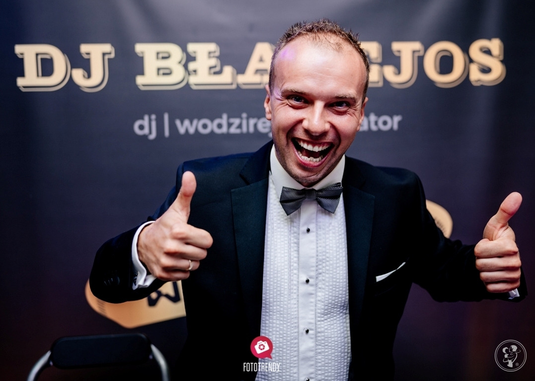 Uśmiechnięty mężczyzna w smokingu i muszce, z uniesionymi kciukami na tle plakatu 'DJ BŁAŻEJOS' i logotypów 'FOTOTRENDY'.