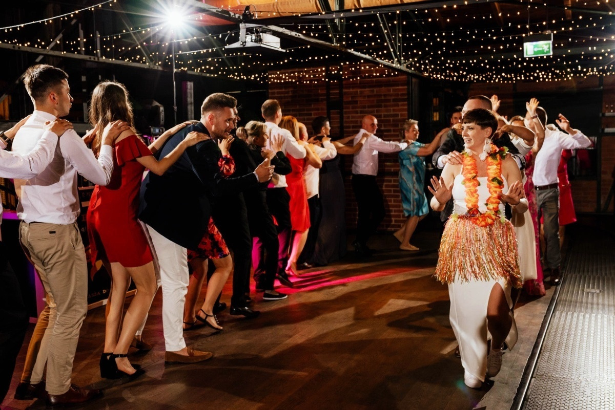 Uczestnicy wesela tańczą w korowodzie, prowadzi kobieta w białej sukni z hawajskim wieńcem i spódniczką z frędzli, oświetlenie dekoracyjne w tle.
