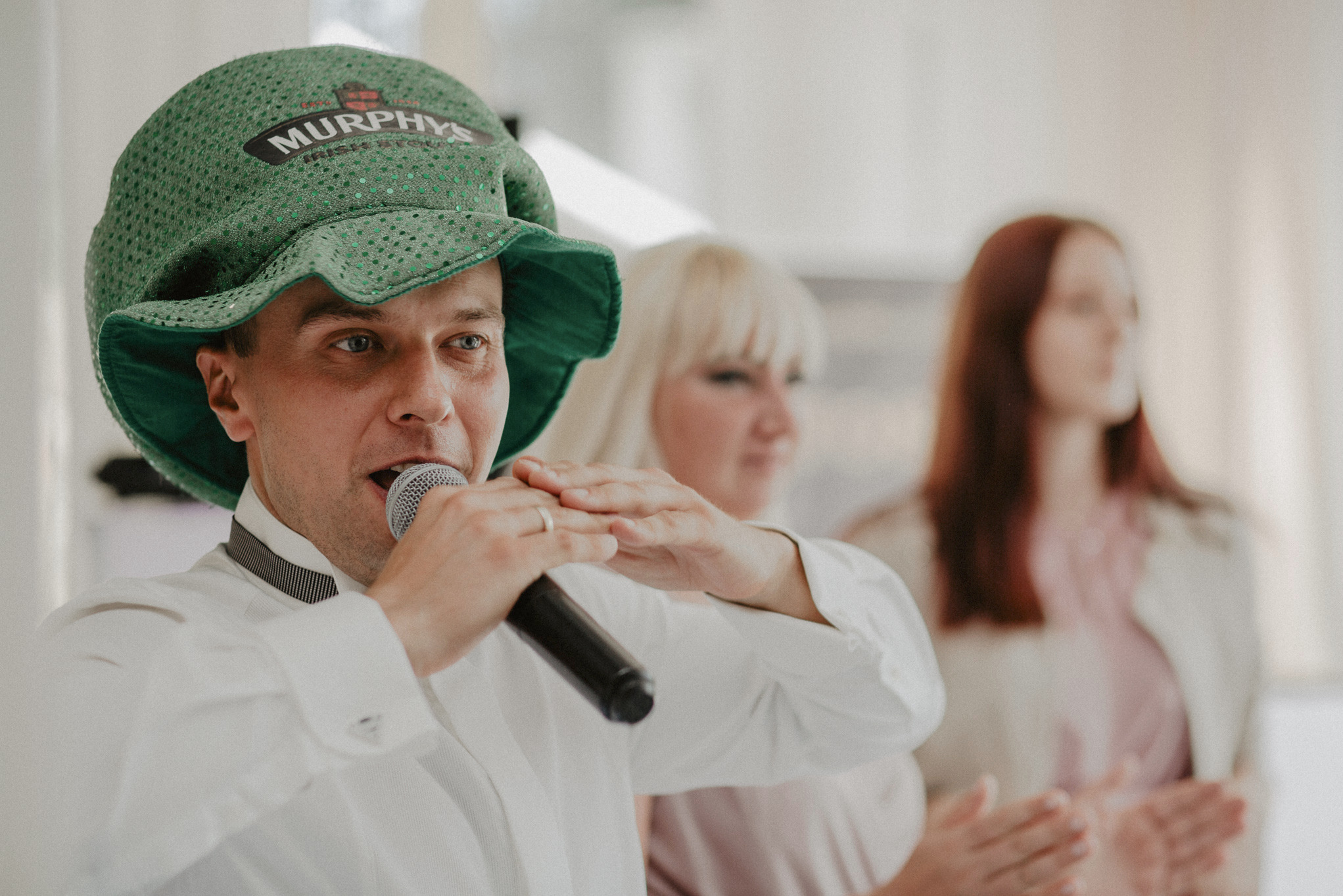 Mężczyzna w zielonym kapeluszu Murphy's śpiewa do mikrofonu, z dłońmi ułożonymi gestykulacyjnie, w tle dwie kobiety.