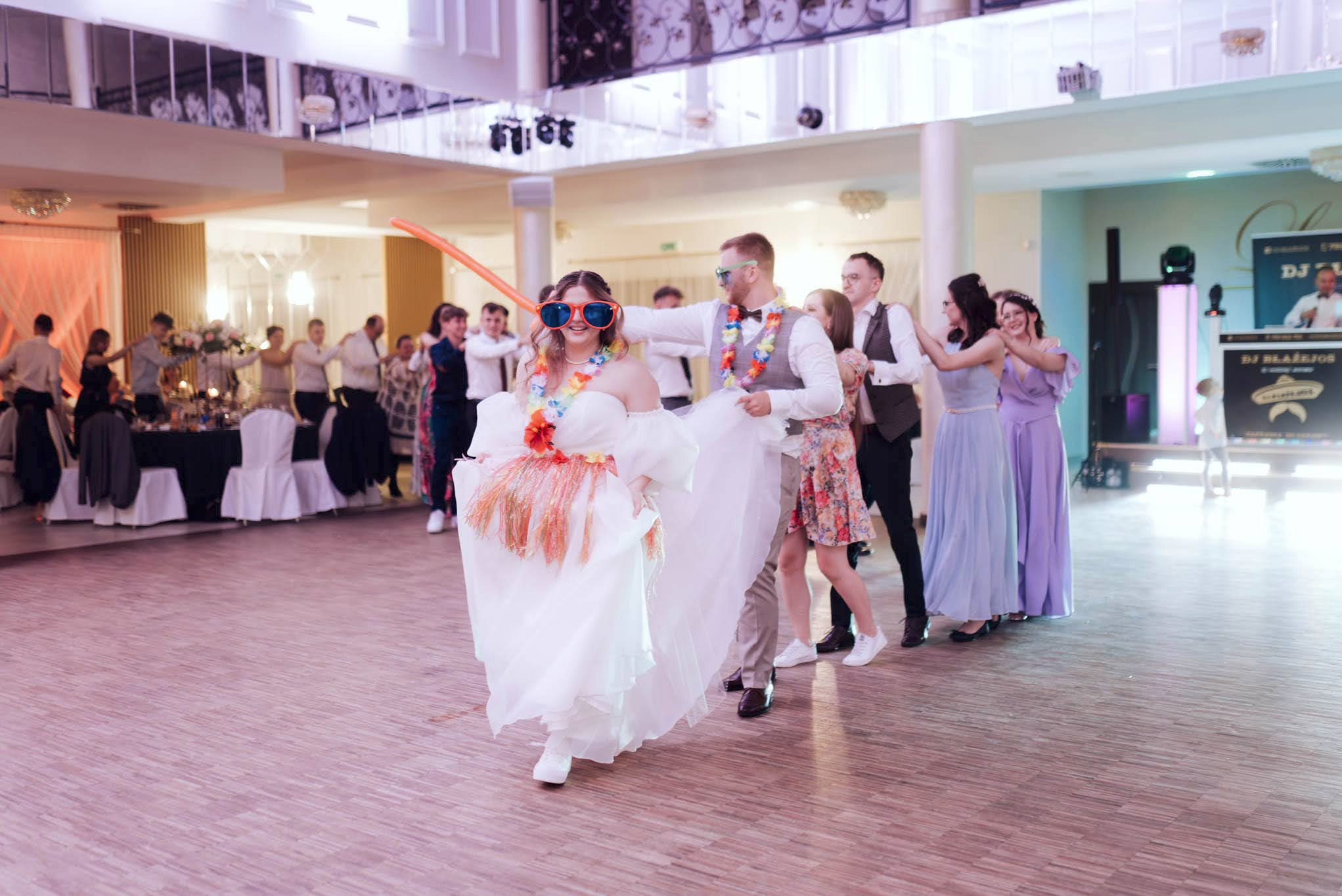 Para młoda w okularach i hawajskich strojach prowadzi taneczny korowód gości weselnych na sali balowej, w tle widoczny DJ.