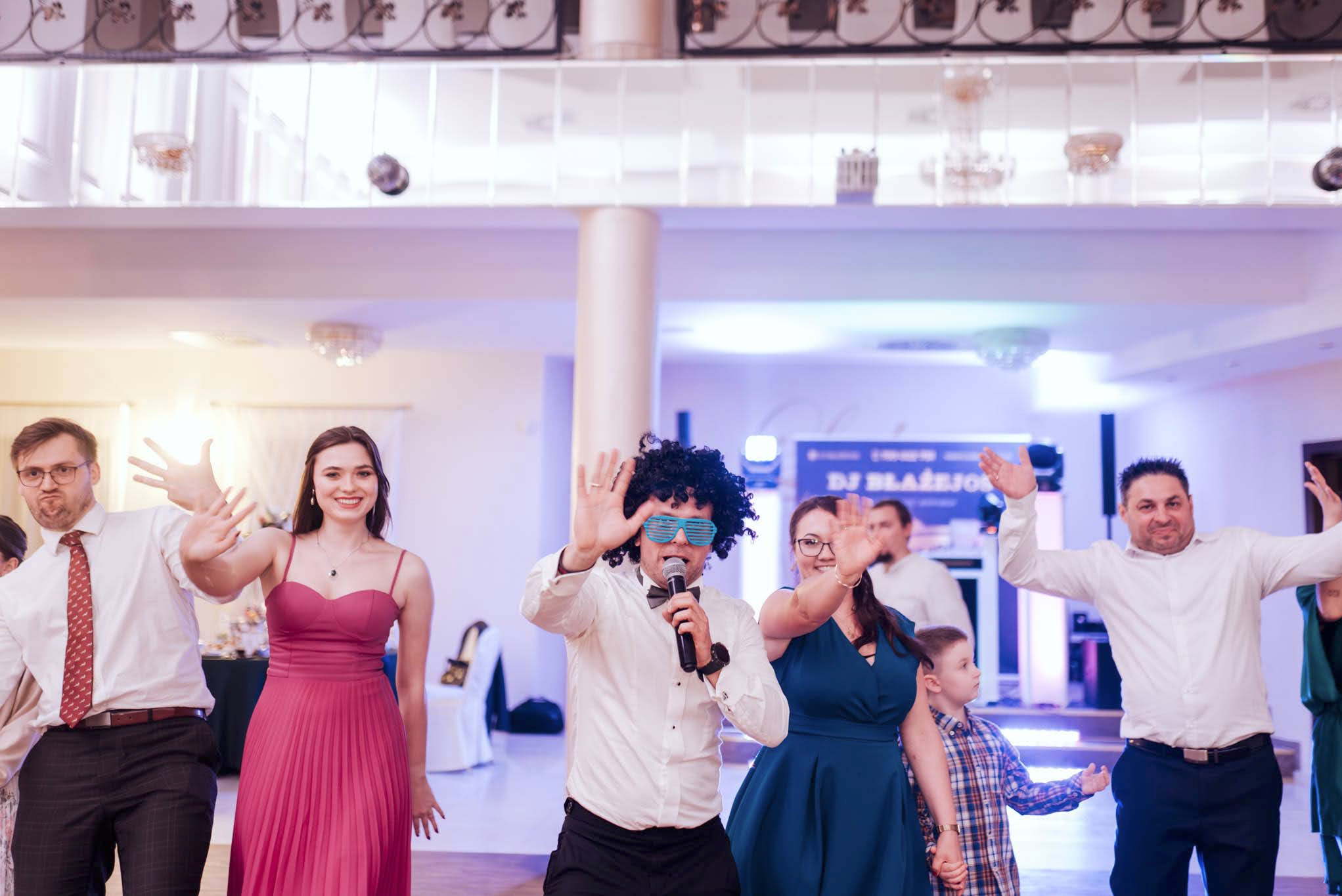 Wodzirej w czarnej peruce i niebieskich okularach, z mikrofonem w ręku, prowadzi zabawę z gośćmi weselnymi na sali balowej z kolumnami i dekoracjami sufitowymi.