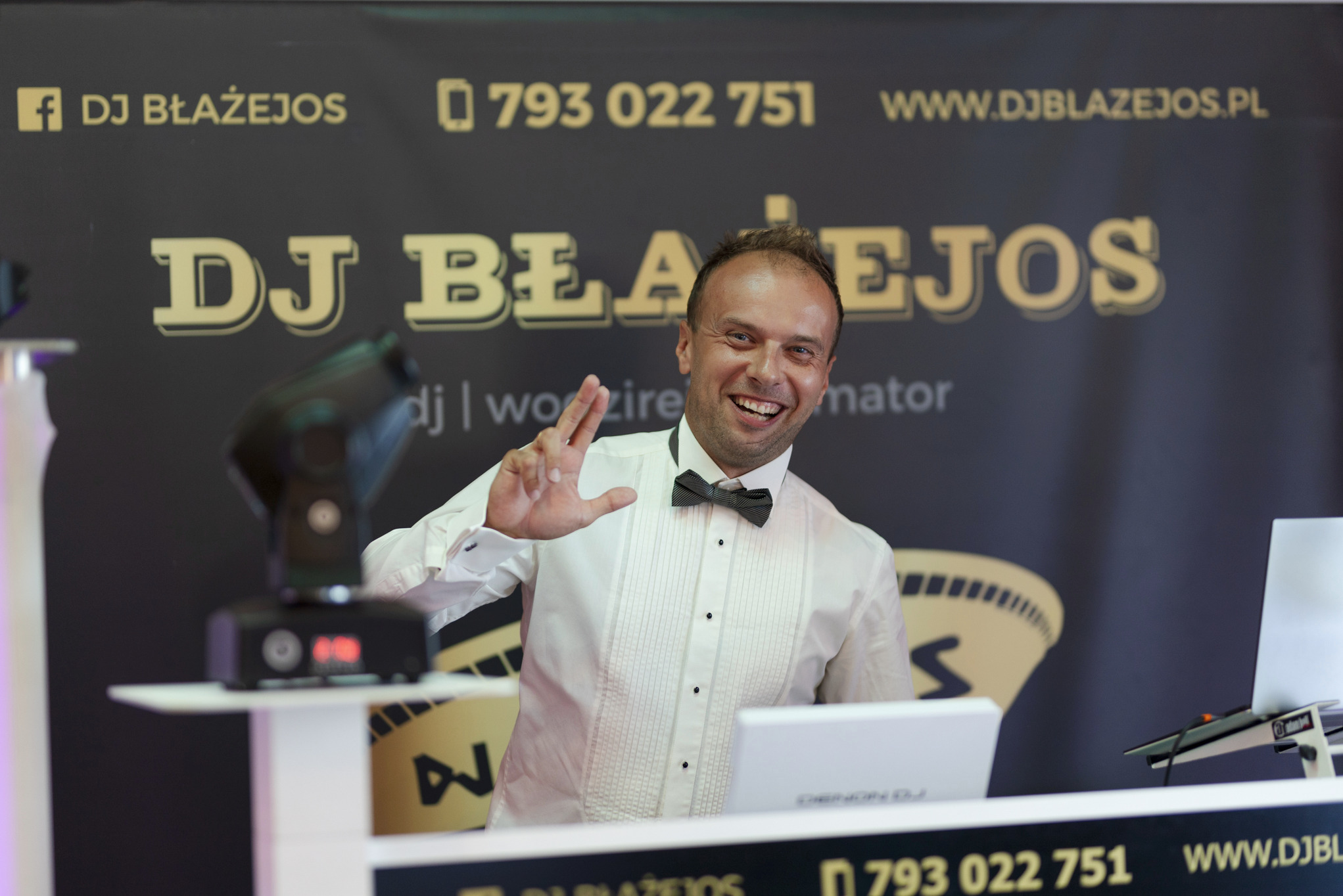 Uśmiechnięty prezenter w muszce, na tle baneru z nazwą DJ Błażejos, w trakcie występu, oświetlenie sceniczne.