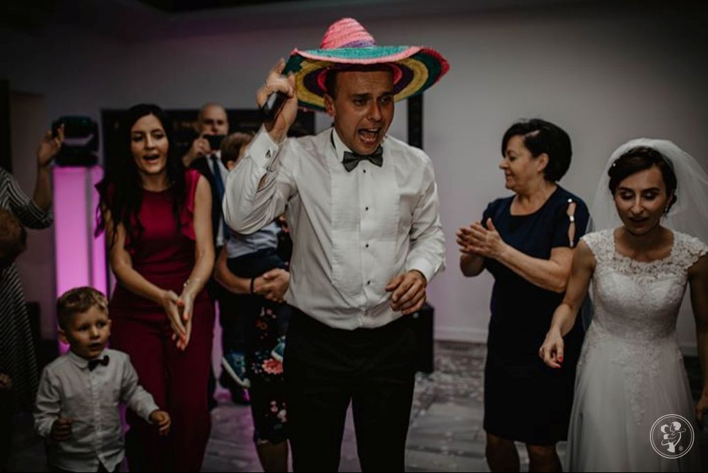 Wokalista w kolorowym sombrero śpiewa do mikrofonu na weselu, obok tańcząca para młoda i goście.