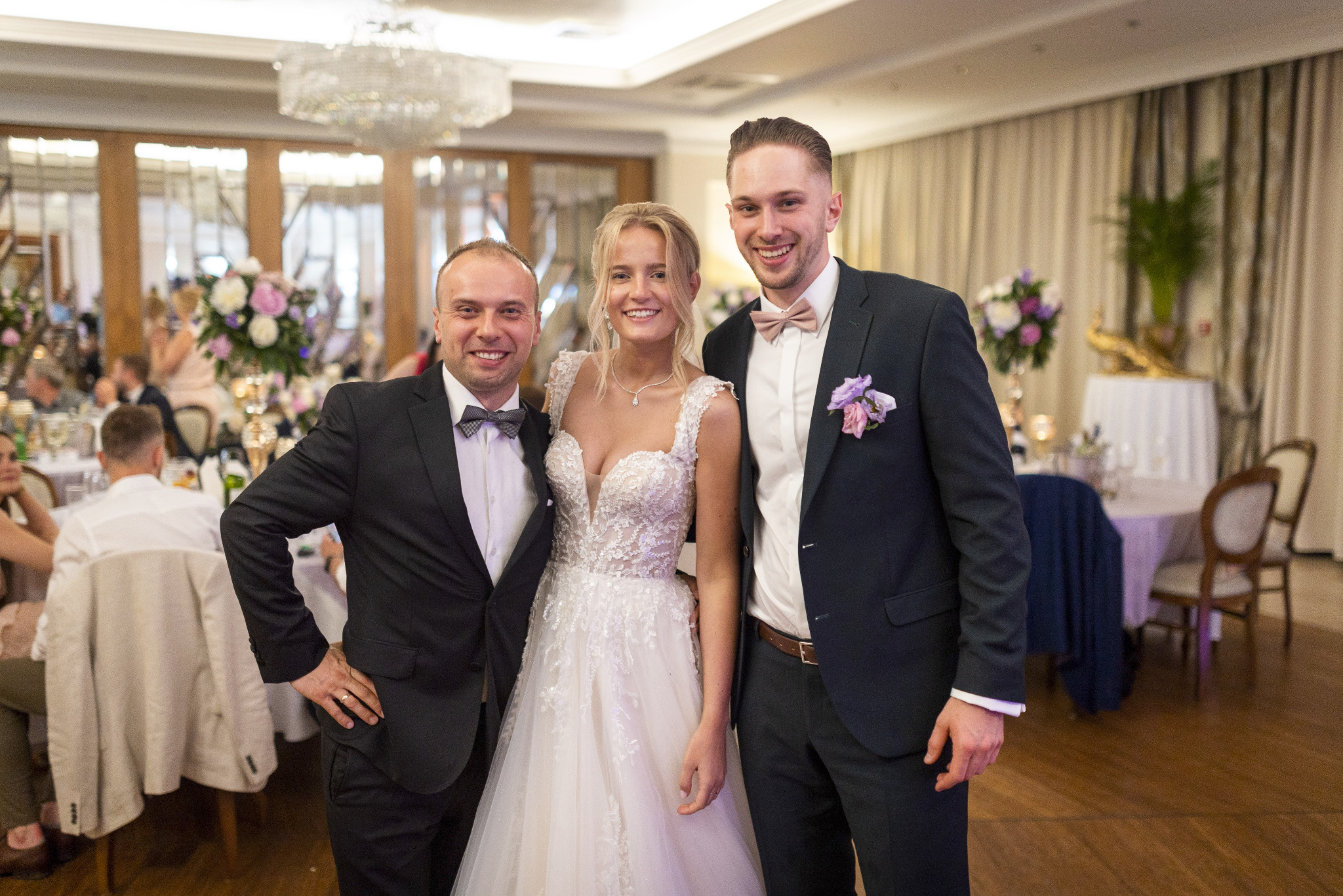 Panna młoda w koronkowej sukni pozuje z dwoma mężczyznami w smokingach na tle sali weselnej z dekoracjami kwiatowymi i kryształowym żyrandolem.