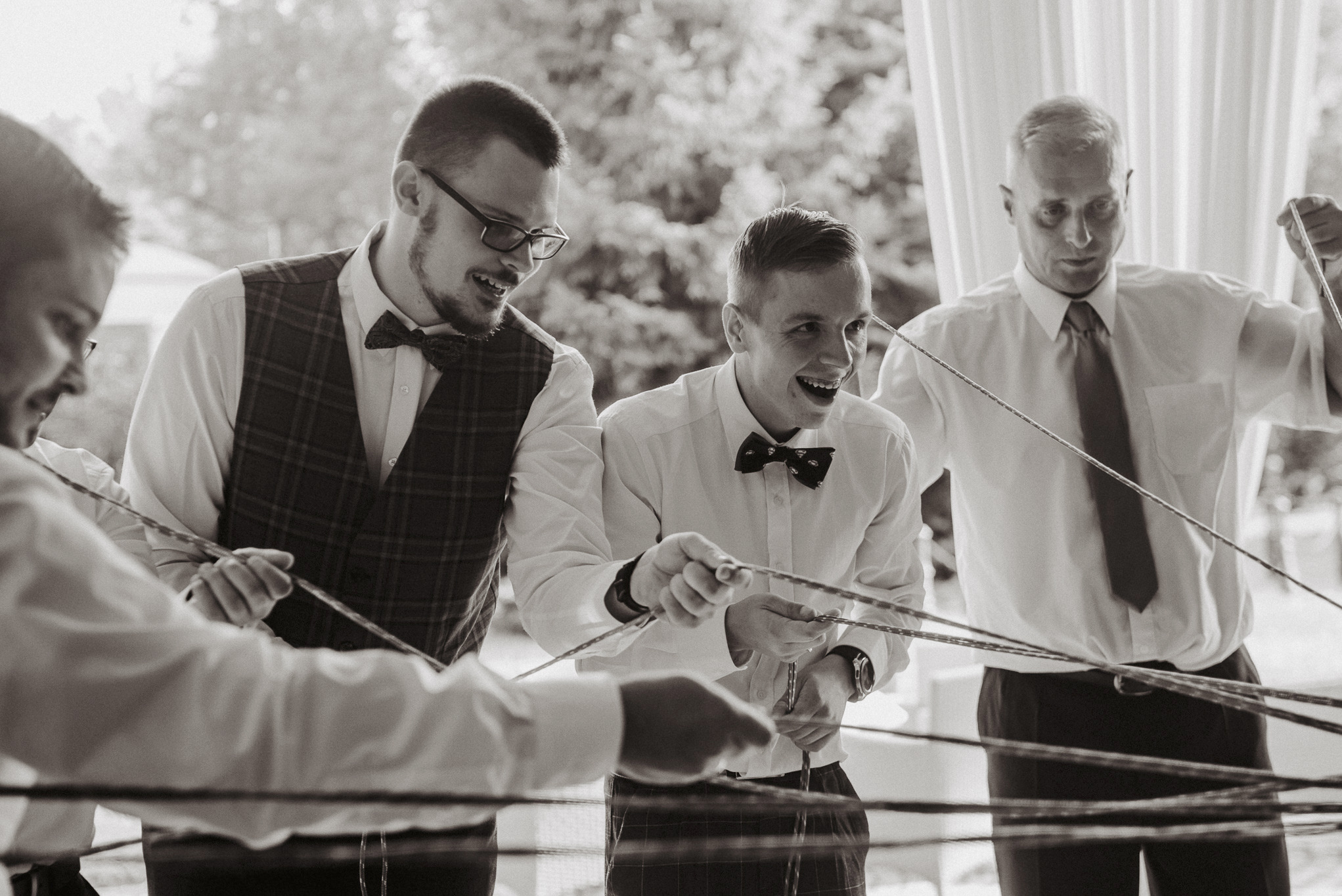 Czarno-białe zdjęcie przedstawia grupę mężczyzn w koszulach i muchach, uśmiechniętych, podczas wykonywania gry zespołowej z wykorzystaniem sznurków na tle białych zasłon, prawdopodobnie podczas...