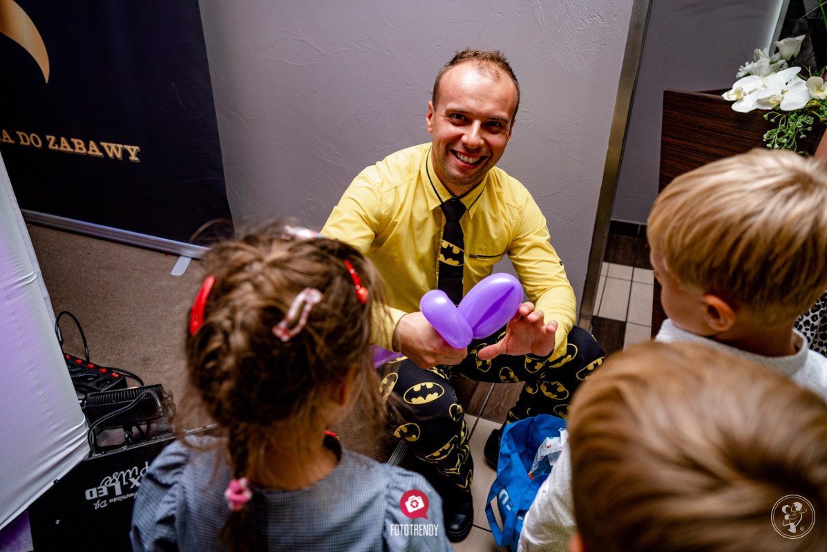 Animator w żółtej koszuli i spodniach z motywem Batmana, uśmiechnięty, modeluje baloniki dla dzieci na imprezie.