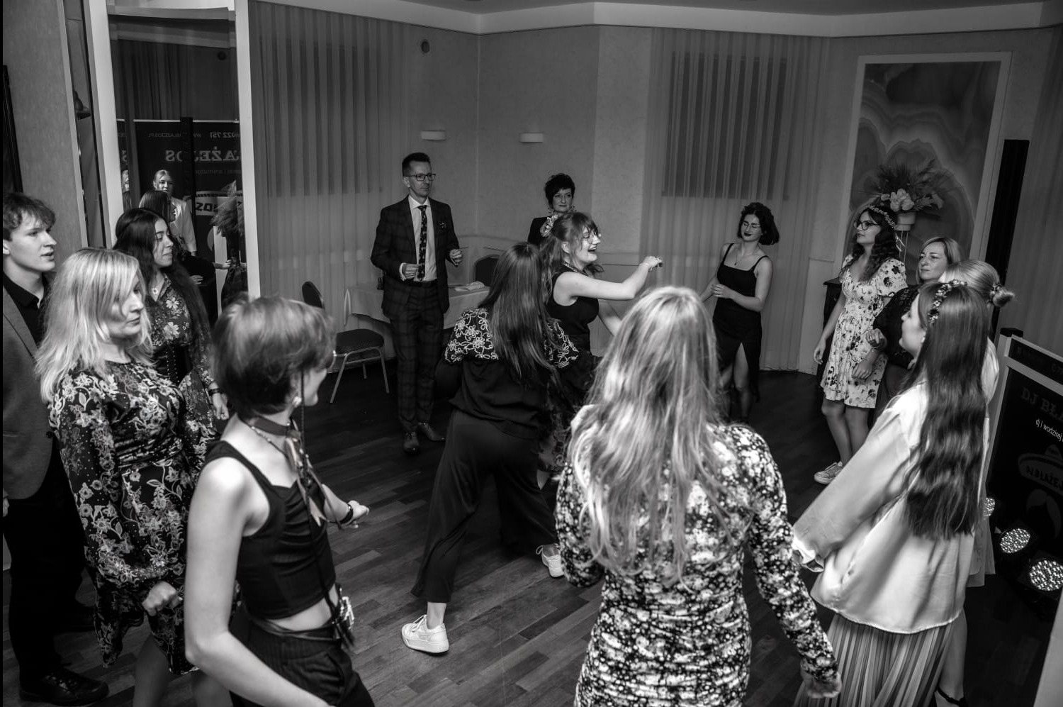 Czarno-białe zdjęcie z imprezy: tańczący goście w kwiecistych sukienkach i garniturach na parkiecie, w tle DJ-ka i dekoracje.