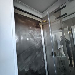 Gatkowski Łukasz - Wykończona kabina prysznicowa z deszczownicą i szklanymi drzwiami, ściana wyłożona płytami imitującymi marmur w odcieniach szarości i brązu, widoczne odbicie osoby w szkle.