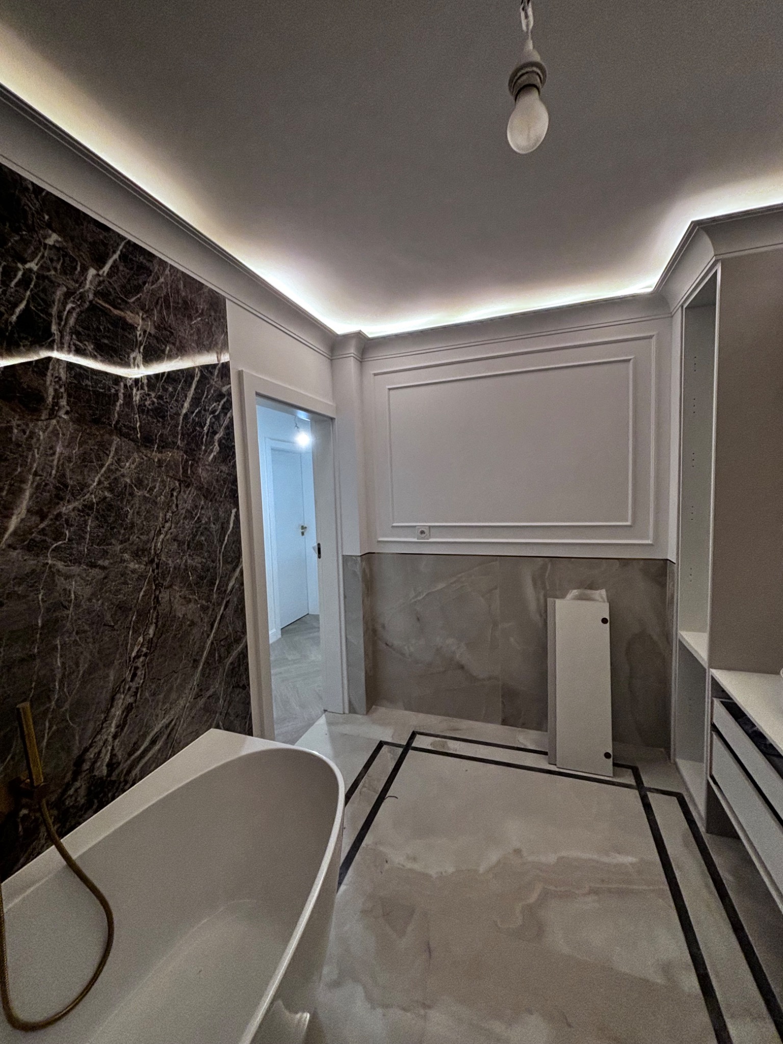 Elegancka łazienka z marmurową ścianą, podświetleniem LED w suficie, białą wanną z złotą baterią i marmurową podłogą z czarnymi akcentami.