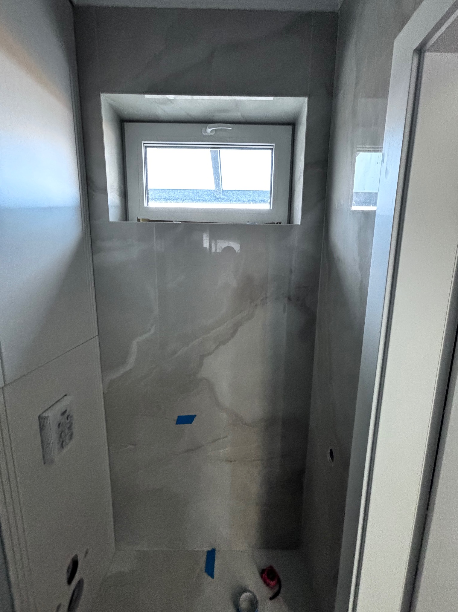 Wnętrze łazienki w trakcie prac wykończeniowych, widoczne płytki imitujące marmur, okno wnękowe, niebieska taśma malarska i narzędzia na podłodze.