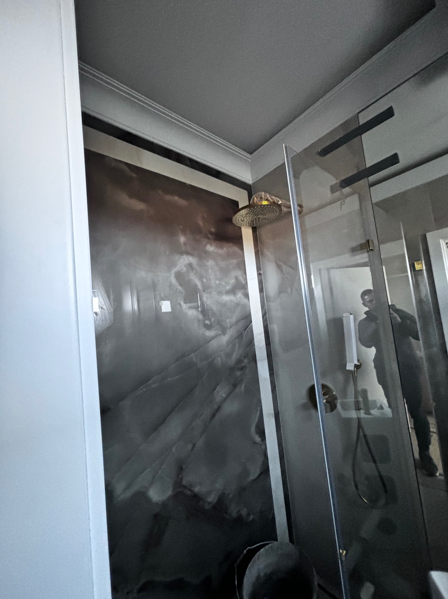 Wykończona kabina prysznicowa z deszczownicą i szklanymi drzwiami, ściana wyłożona płytami imitującymi marmur w odcieniach szarości i brązu, widoczne odbicie osoby w szkle.