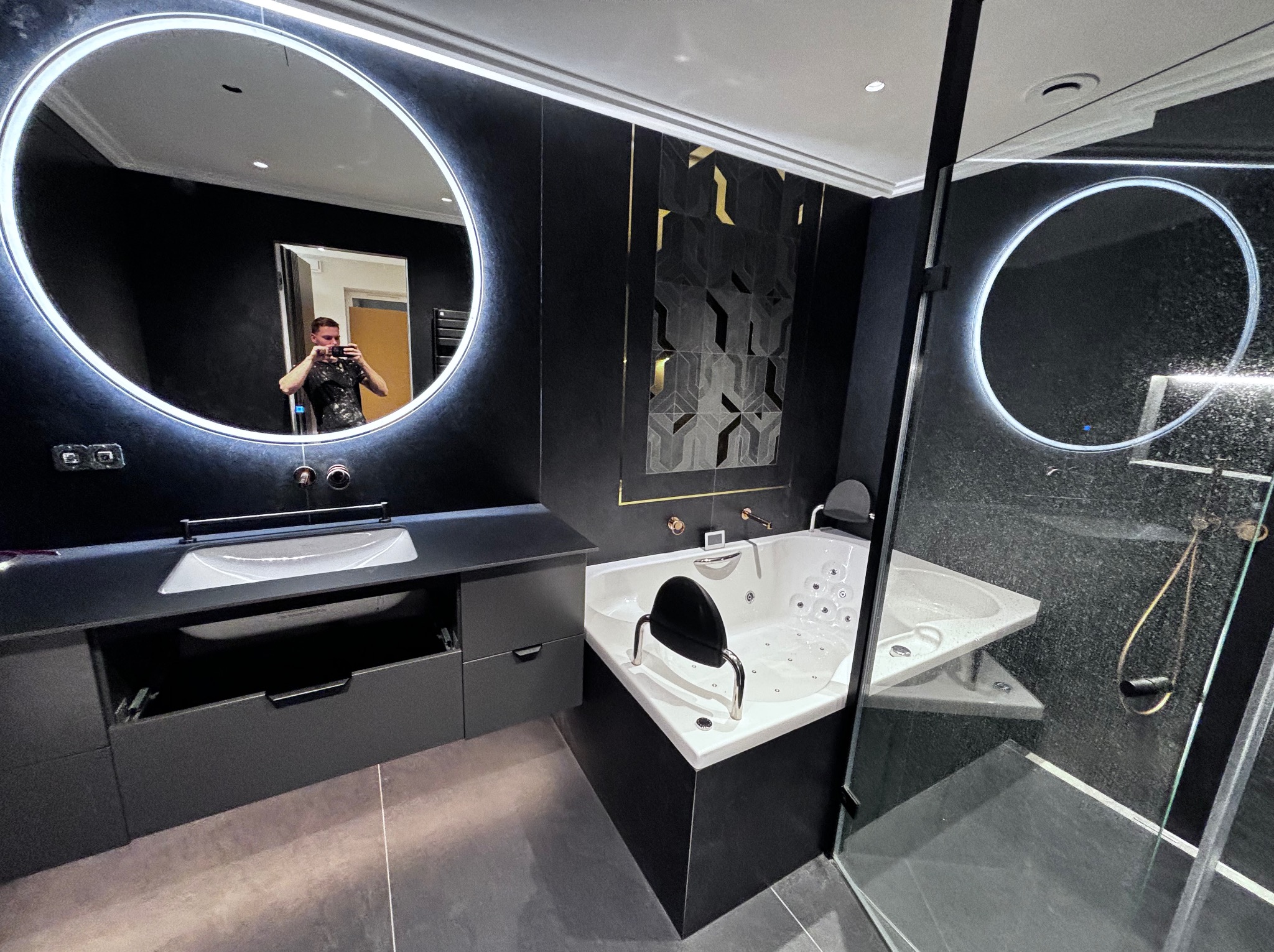 Nowoczesna łazienka w ciemnej kolorystyce z podświetlanymi lustrami, narożną wanną z hydromasażem i kabiną prysznicową ze szklanymi drzwiami, widoczne odbicie osoby robiącej zdjęcie w lustrze...